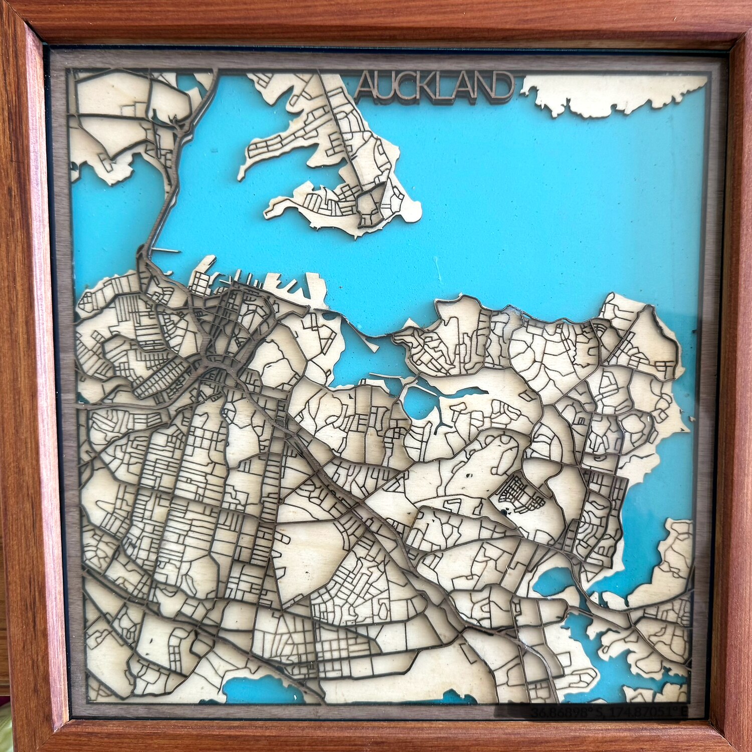 Layered laser-cut relief map of Auckland: native ply suburbs stacked in relief with the harbour poured in blue resin, in a timber shadow box