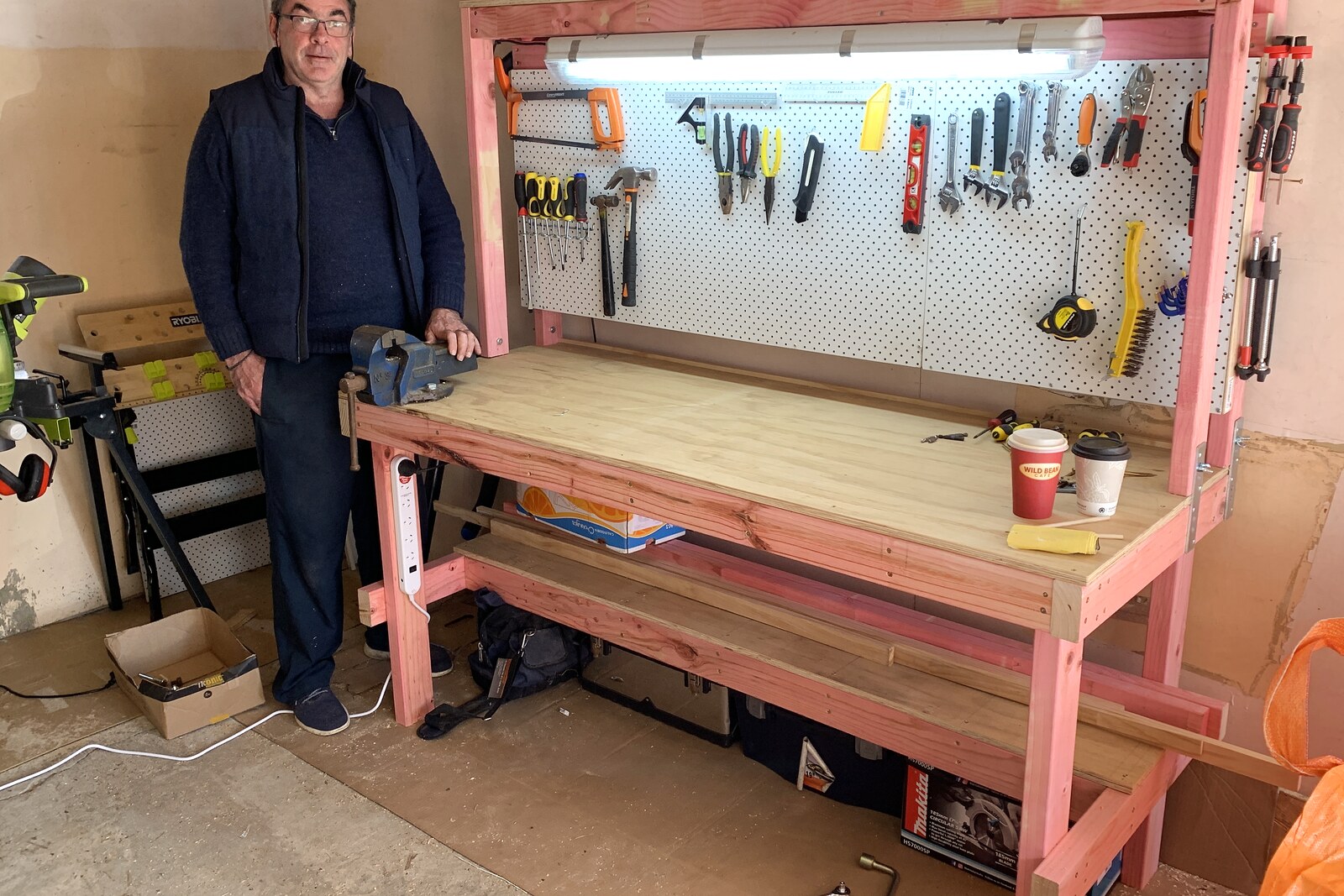 Mark in his garage workshop, standing at the workbench he built, tools on a pegboard behind him