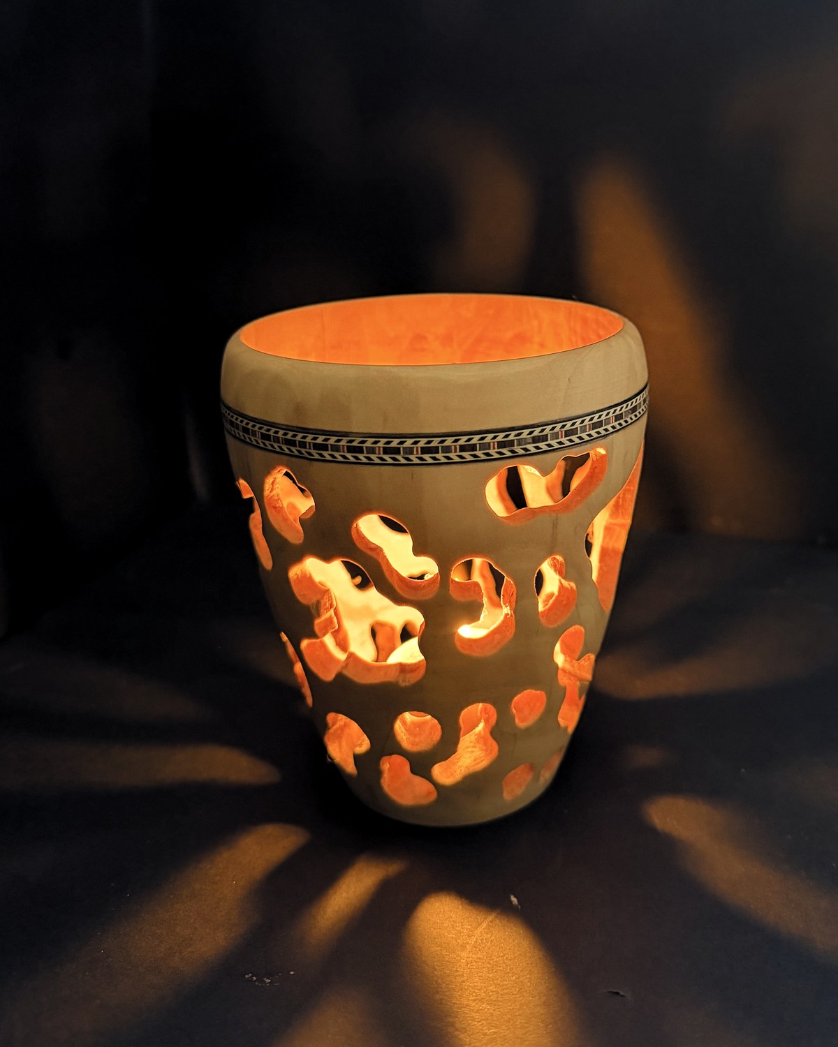 Pierced turned vessel lit from within, the openings glowing and throwing their pattern onto the wall behind, with an inlaid band at the shoulder
